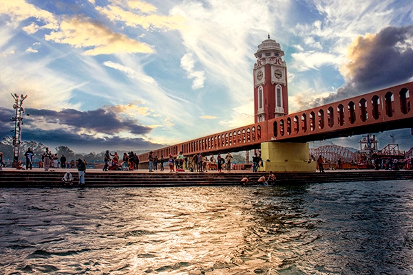 Scenic view of Haridwar with people by the Ganges and an ornate bridge with a tower.