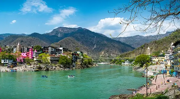 Scenic view of Rishikesh with Ganges river, colorful buildings, and mountain backdrop.
